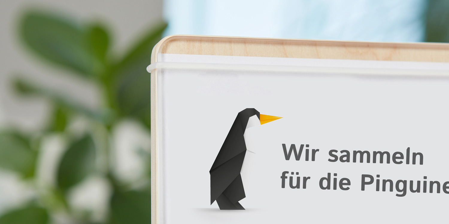 Die Displayfläche ist eine Holzplatte, auf die das selbst gestaltete A4-Blatt gelegt werden kann. Darauf wird eine transparente Acrylscheibe gelegt und mit einem Silikonring befestigt. Der Spendenaufruf zeigt: Wir sammeln für die Pinguine!